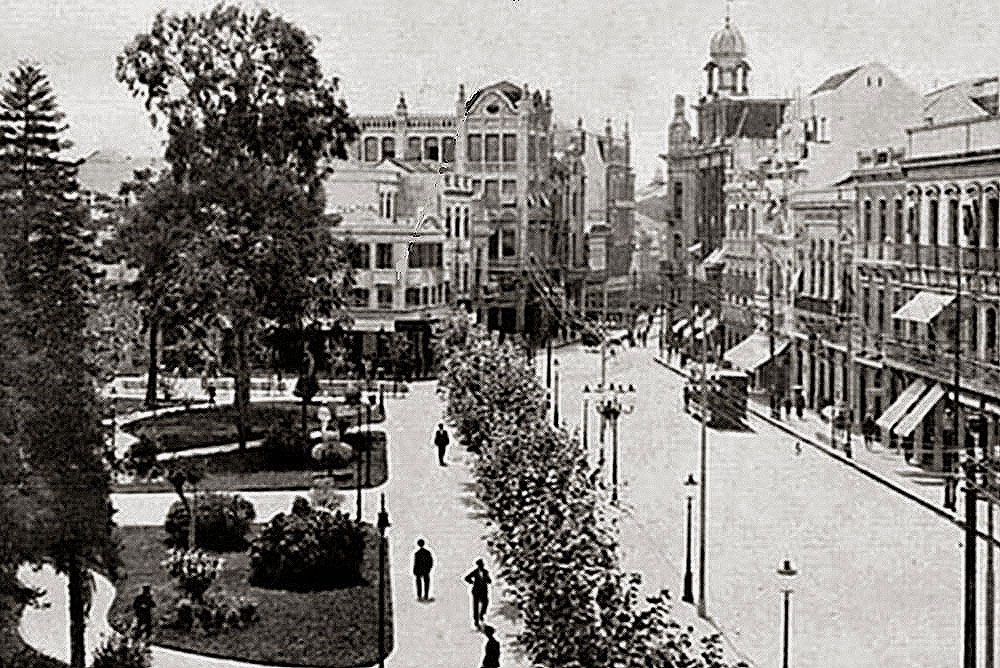 Fotografia em preto e branco do centro de Porto Alegre por volta de 1900: praça arborizada, sobrados e um bonde na rua.