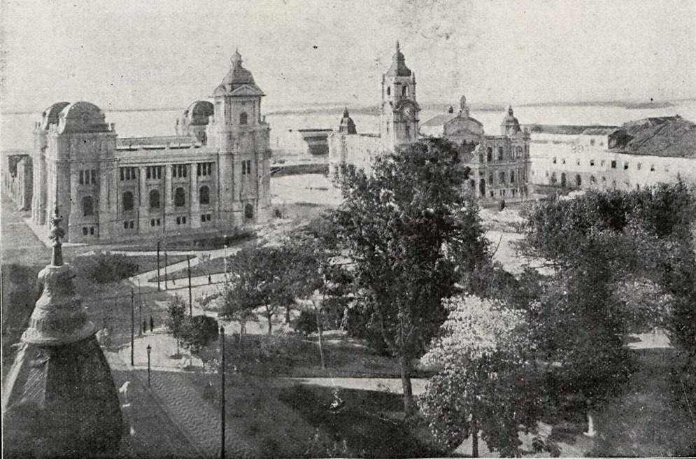 Fotografia antiga da Praça da Alfândega, em Porto Alegre, em 1919: árvores da praça, os prédios dos Correios e da Delegacia Fiscal e o Guaíba ao fundo.