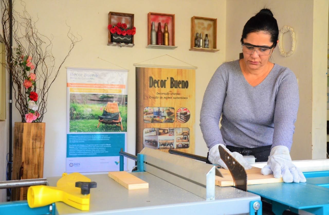 Simone Bueno cortando madeira na serra de mesa, com óculos de proteção e luvas, na oficina da Decor Bueno