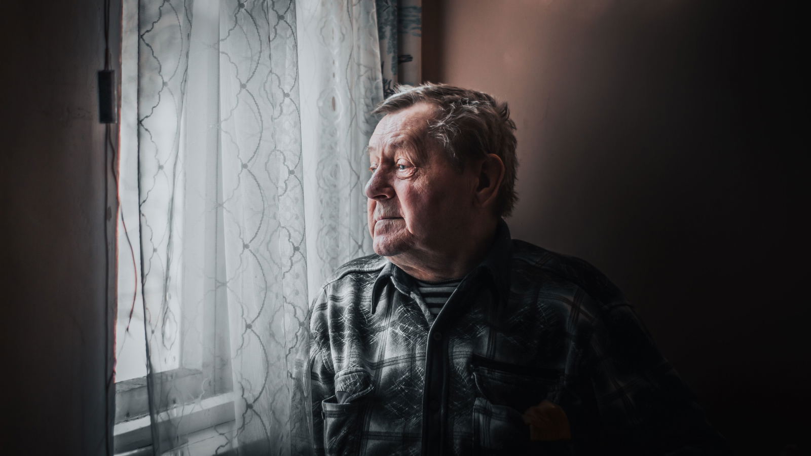 Idős férfi elgondolkodva néz ki az ablakon otthonában